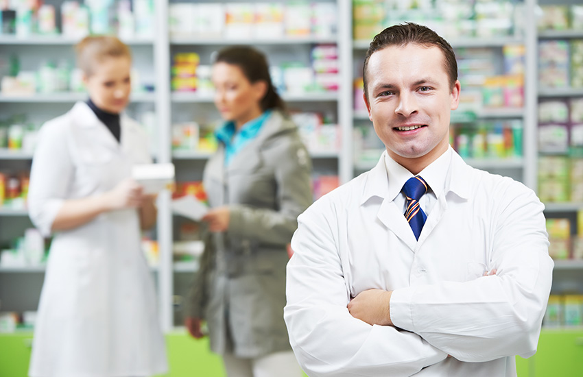 Farmac&eacute;utico en primer plano con los brazos cruzados sonr&iacute;e mientras en el fondo una farmac&eacute;utica ayuda a una clienta con un producto