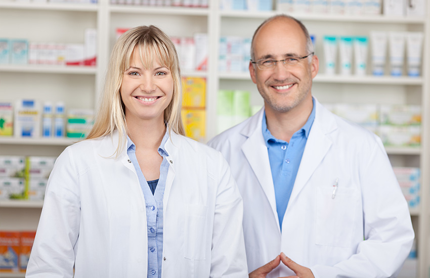 Dos farmac&eacute;uticos sonr&iacute;en mirando al frente con estanter&iacute;a de productos detr&aacute;s
